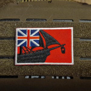 May include: A red and white embroidered patch with a black silhouette of a ship and the Union Jack flag. The patch is sewn onto a brown fabric background.