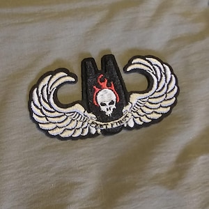 May include: Embroidered patch featuring a skull with flames, set within a black and white winged design. The patch has the text "FIRST TO FIGHT" and is likely for a jacket or bag.