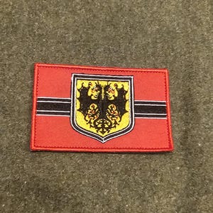 Könnte beinhalten: Ein rechteckiger roter Aufnäher mit einem schwarzen horizontalen Streifen und einem zentralen Emblem. Das Emblem zeigt einen gelben Schild mit einem schwarz-goldenen Design. Der Aufnäher ist wahrscheinlich ein Emblem oder ein Abzeichen.
