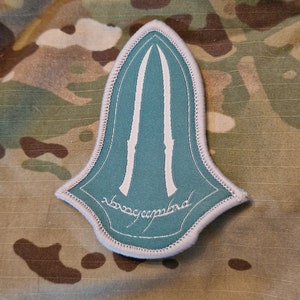 Puede incluir: Un parche bordado verde y blanco con forma de punta de lanza con el texto "The One Ring" en una fuente cursiva.