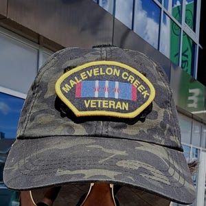 May include: Camouflage baseball cap with a patch that reads "Malevelon Creek Veteran". The patch has a yellow border and features a design with red, white, and blue stripes and three white symbols.