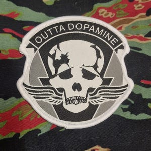 May include: A white, black, and gray patch with the text "OUTTA DOPAMINE" encircling a skull with wings. The patch is on a camouflage background.