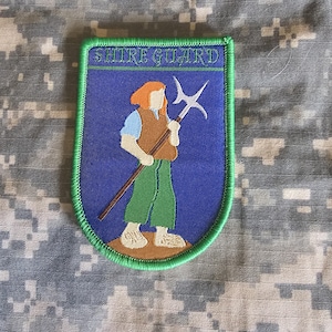 Op de afbeelding: Geborduurde patch met de tekst "SHIRE GUARD" bovenaan. De patch toont een figuur met een hellebaard, met oranje haar, een bruin vest, groene broek en een blauwe achtergrond. De patch heeft een groene rand en staat op een camouflage achtergrond.