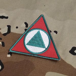 Può includere: Una toppa ricamata triangolare con sfondo rosso e bordo verde acqua. Al centro, un cerchio bianco contiene un triangolo verde acqua pieno. La toppa è cucita con un motivo a filo denso, creando una superficie strutturata su tutto il design.