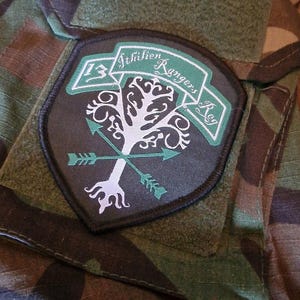 Könnte beinhalten: Ein Aufnäher mit dem Text "13 Ithilien Rangers Reg" und einem weißen Baumdesign mit gekreuzten grünen Pfeilen. Der Aufnäher befindet sich auf einem Tarnhintergrund.