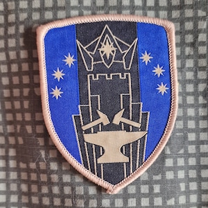 Könnte beinhalten: Ein blauer und brauner gestickter Aufnäher mit einem Schloss, Sternen und einem Amboss. Das Schloss hat eine Krone oben und der Amboss hat zwei Hämmer oben.