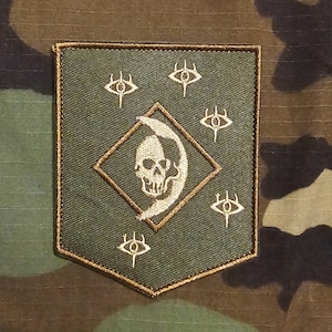 Puede incluir: Un parche verde y marrón con un diseño de calavera y media luna dentro de un rombo, rodeado de símbolos de ojo. El parche tiene un borde marrón y está sobre un fondo de camuflaje.