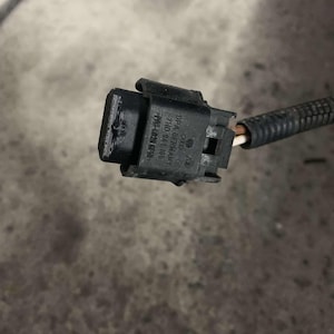 Op de afbeelding: Een zwarte elektrische connector met een grijze draad eraan bevestigd. De connector heeft de tekst "VW AG" en "MADE IN GERMANY" erop gedrukt.