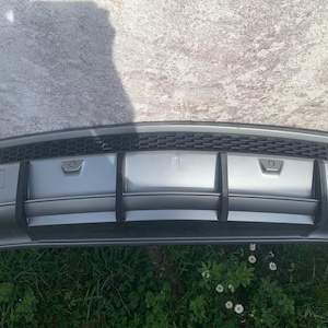 Può includere: Un paraurti posteriore per auto argentato con una griglia a rete nera e quattro alette nere. Il paraurti ha una superficie testurizzata ed è progettato per migliorare l'aspetto dell'auto.