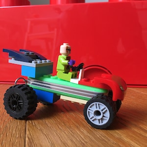 Könnte beinhalten: Ein grünes und rotes Lego-Auto mit einem blauen und grauen Flügel, eine grüne Lego-Figur mit Glatze und lila Handschuhen fährt das Auto.