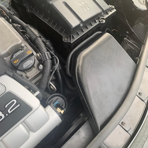 May include: A car engine bay featuring a black air intake box and other parts. The engine block is silver, displaying "3.2". Hoses and wires are present, along with a black air filter housing.