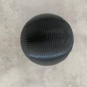 Peut inclure: Une sphère noire avec une surface texturée et striée. L'objet a un sommet lisse et arrondi et est posé sur un fond gris. Le diamètre de la sphère est d'environ 7 cm.
