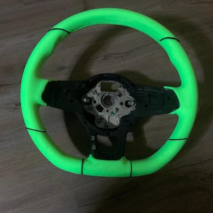 May include: A vibrant neon green steering wheel with black detailing. The wheel features a circular design with a black central component and black stitching. It is set against a wooden surface.