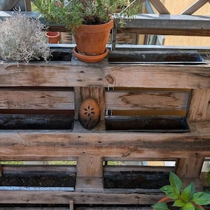 Könnte beinhalten: Ein rustikaler Holzpaletten-Pflanzkasten mit mehreren rechteckigen Metallbehältern für Pflanzen. Der Pflanzkasten enthält verschiedene Pflanzen, darunter Rosmarin und anderes Grün. Ein kleines dekoratives Holzstück mit Blumenmuster ist befestigt.