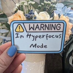 Op de afbeelding: Een blauwe en gele waarschuwingssticker met de tekst "WARNING In Hyperfocus Mode".