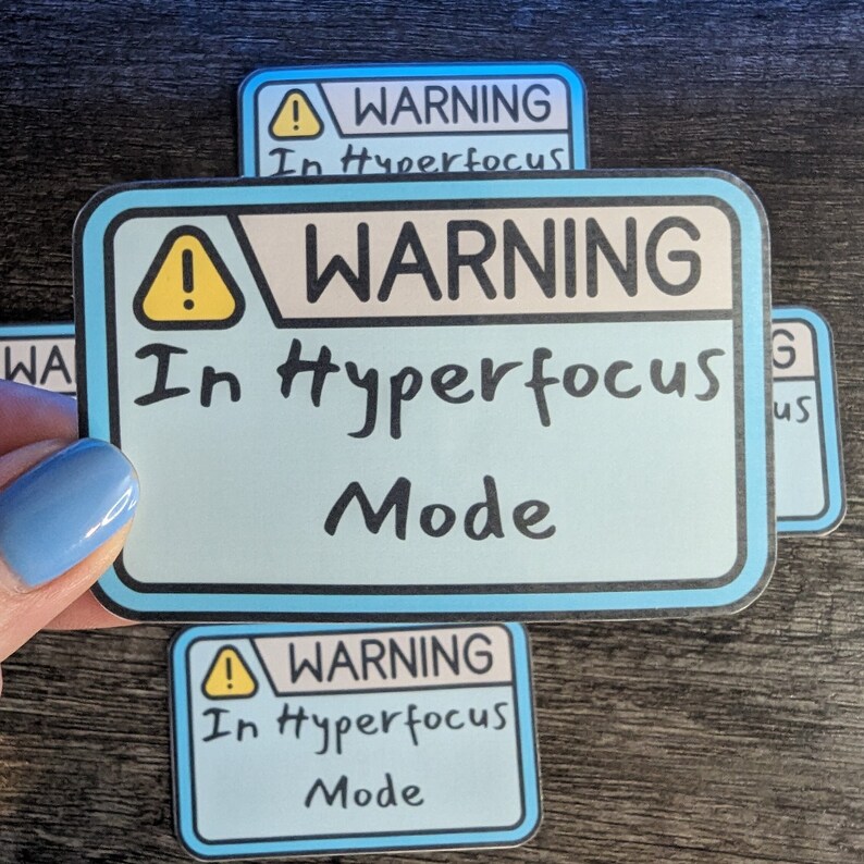 以下が含まれることがあります： 木製の表面に、「WARNING In Hyperfocus Mode」と書かれた青と黄色の警告ステッカーが3つあります。