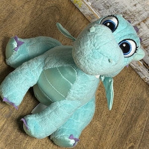 Puede incluir: Un peluche de dragón turquesa con ojos grandes y expresivos y detalles morados en garras y espinas. El dragón tiene un pelaje suave y texturizado y una barriga satinada. Las alas son pequeñas y delicadas.