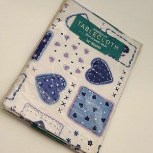 May include: A white vinyl flannel back tablecloth in a 60-inch round size, packaged in clear plastic. The fabric features a blue and lavender heart pattern, decorative borders, and small floral accents. A teal label reads Home Trends Tablecloth.