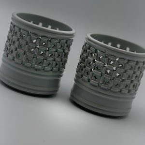 May include: Two grey ceramic candle holders. Each cylindrical holder features a geometric pattern of cut-out shapes around the body. The top rim has small circular openings. The holders are a muted grey colour.