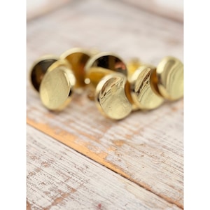 May include: A set of small, circular, gold-colored metal buttons with a smooth, reflective surface. Each button features a slightly domed, convex shape and a polished finish that catches the light, creating bright highlights across the metallic surface.