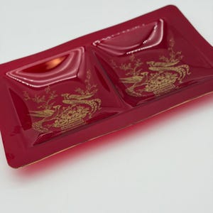 May include: A rectangular, deep red glass divided tray featuring two square compartments. Each compartment is decorated with a gold-colored illustration of a bird perched on a flowering branch. The edges of the tray are trimmed in gold.