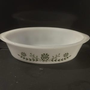 May include: White oval casserole dish with a green floral design. The dish has a smooth, glossy finish and is decorated with stylized flowers and leaves. The dish is likely made of glass or ceramic and is suitable for baking or serving.