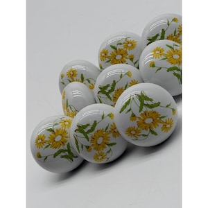 May include: A collection of nine round white ceramic cabinet knobs, each featuring a painted pattern of yellow daisy flowers with green stems and leaves. The knobs are arranged in a cluster, showcasing their smooth, glossy, and spherical surface.