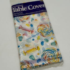 May include: A rectangular plastic table cover in its original packaging. The cover features a white background with a colorful pattern of swirled lollipops, wrapped candies, and jelly beans. The label states it measures 54 inches by 96 inches.