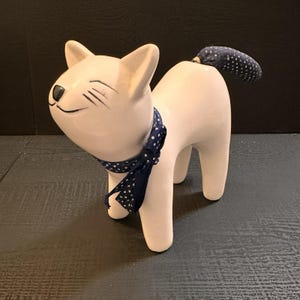 May include: A white ceramic cat ornament with a navy blue polka dot scarf and tail. The cat has a smiling face with black details. The tail is navy blue fabric with white polka dots. The cat stands on four legs.
