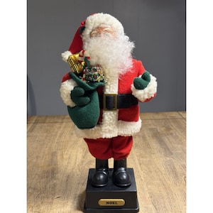 Pode incluir: Uma estatueta do Pai Natal vestida com um fato vermelho com acabamentos em pele sintética branca, um cinto preto e botas pretas. Ele segura um saco verde cheio de presentes. A base da estatueta diz "NOEL".