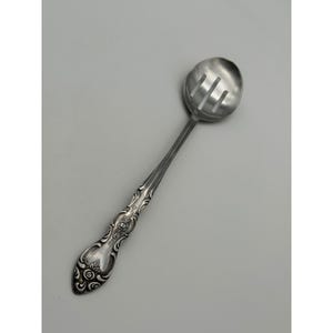 May include: A silver-toned slotted serving spoon featuring a round bowl with three vertical drainage slits. The handle is decorated with an embossed floral and scroll pattern that extends from the neck down to the rounded, tapered end.