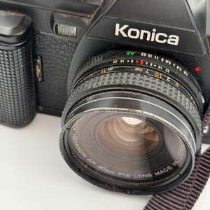 May include: A black Konica SLR camera featuring a Hexanon AR 40mm F1.8 lens. The lens barrel displays aperture settings from 1.8 to 22, distance markings in meters and feet, and the text Konica Hexanon AR 40mm F1.8 Lens Made in Japan.