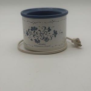 May include: A white Rival Potpourri Crock electric warmer featuring a blue floral pattern and a blue rim. The cylindrical ceramic device includes an attached white power cord with a standard two-prong plug, designed for heating aromatic potpourri.