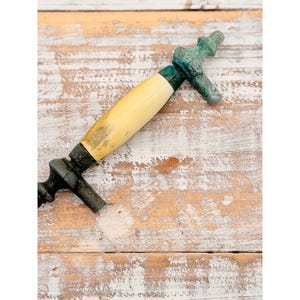 May include: A drawer pull featuring a central cream-colored cylindrical handle flanked by two dark, weathered metal ends with a textured, oxidized green patina. The hardware displays a classic shape with decorative finials at each mounting point.