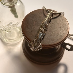 May include: A silver pendant necklace with a spoon-shaped design, featuring grape cluster detailing. The pendant hangs on a silver chain and rests on a wooden stand. A clear glass bottle is in the background.