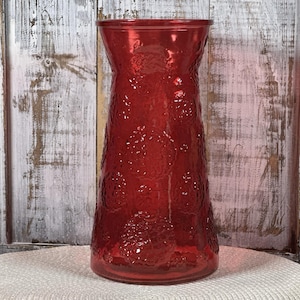May include: A tall, red glass vase with a textured surface and a flared top. The vase features a repeating pattern of raised circular designs. The background is a distressed white wooden surface.