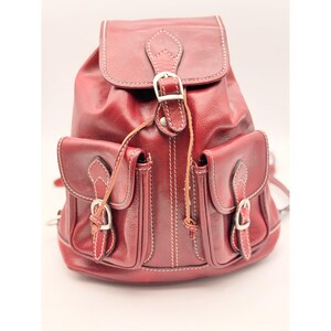 May include: A deep red leather backpack featuring a top flap with a silver buckle closure, two front pockets with matching silver buckles, and white contrast stitching throughout. The bag includes a drawstring closure and adjustable shoulder straps.