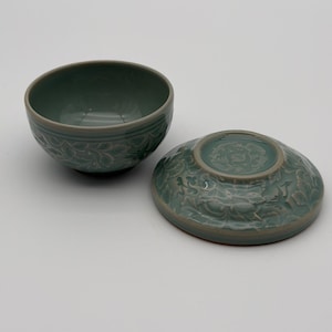 May include: A celadon ceramic bowl and lid. The bowl has a detailed floral relief pattern around the rim. The lid, also celadon, features a similar design. The set is displayed on a white surface.