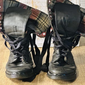 Puede incluir: Par de botas de combate de cuero negro con forro de tela a cuadros. Las botas tienen cordones y una parte superior doblada. La tela a cuadros presenta colores rojos, marrones y verdes. Las botas están sobre una superficie de madera.
