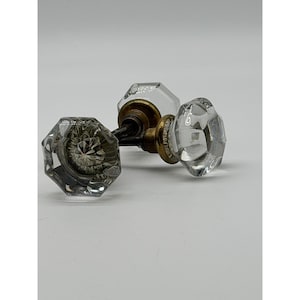 May include: Two octagonal clear glass door knobs featuring faceted surfaces that catch the light. Each knob is attached to a circular brass-toned metal base with a threaded metal spindle extending from the center, connecting the two pieces together.