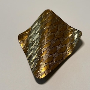Può includere: Spilla vintage a forma di diamante con superficie strutturata. La spilla presenta un motivo di forme a foglia nei toni dell'oro e dell'argento, creando un effetto dimensionale a strati. Il metallo ha una leggera onda.