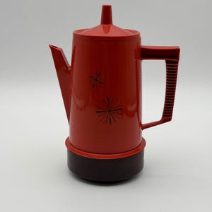 May include: A vintage red coffee pot with a dark brown base and handle. The pot features a lid and spout, with two black starburst designs on the front. The handle has vertical ridges.