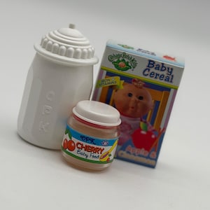 May include: A white plastic bottle with the letters CPK embossed on the side, a small jar of cherry baby food with a white lid, and a rectangular box of Cabbage Patch Kids Apple and Cinnamon baby cereal featuring a doll illustration.