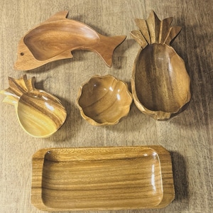 Puede incluir: Una colección de cuencos y bandejas de madera para servir en varias formas. Incluye un pez, una piña y un cuenco festoneado, junto con una bandeja rectangular. Los cuencos y la bandeja son de madera de color marrón claro.