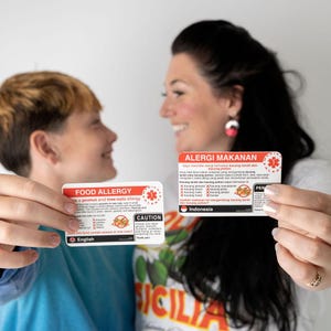 以下が含まれることがあります： 2人の人物が食物アレルギーカードを持っています。カードには、英語で「FOOD ALLERGY」と「CAUTION」と書かれた赤と白の文字があります。もう一方のカードにはインドネシア語で「ALERGI MAKANAN」と書かれています。カードには、ピーナッツとナッツのアレルギーに関する情報が記載されています。