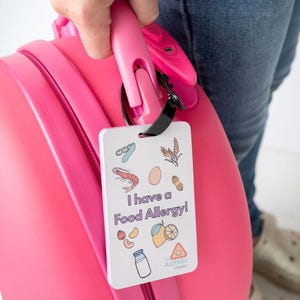 Pode incluir: Uma etiqueta de bagagem branca com o texto "Eu tenho alergia alimentar!" e ilustrações de alérgenos comuns. A etiqueta está presa a uma alça de mala rosa. O logotipo do The Allergy Project está na parte inferior.