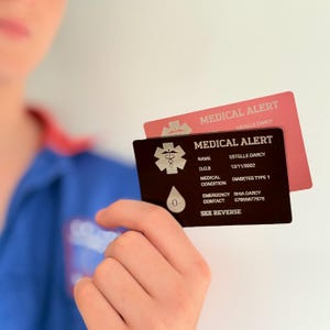 Puede incluir: Dos tarjetas de alerta médica se sostienen frente a una persona. La tarjeta frontal es de color marrón oscuro con texto blanco y un símbolo médico. La tarjeta superior es rosa con las palabras "MEDICAL ALERT". Las tarjetas proporcionan información médica.