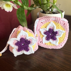 May include: Two handmade crochet items: a small drawstring bag and a square coaster. Both are pink, yellow, white, and purple, featuring a large flower design. The bag has a drawstring closure with a wooden bead. The coaster has a similar flower design.