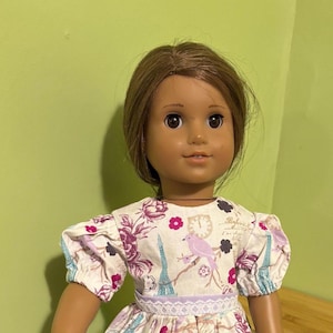 May include: A doll wearing a lavender and white dress with a floral print featuring birds, Eiffel Towers, and a clock. The dress has a lace trim and a lavender ribbon.