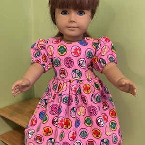 May include: A doll in a pink dress patterned with colourful circular designs. The dress features short, puffed sleeves and a gathered skirt. The doll has brown hair and blue eyes.